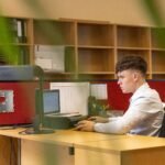 A focused young man typing on a computer in a contemporary office environment.
