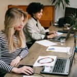 Two women collaborating at their laptops in a modern office setting, fostering teamwork and creativity.